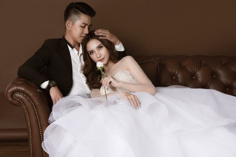 chú rể mặc vest đen ôm hôn đầu cô dâu đang nằm trên ghế sofa