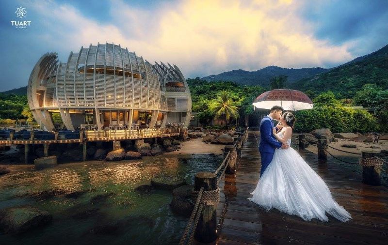 chú rể mặc áo vest xanh dương ôm cô dâu cùng che ô