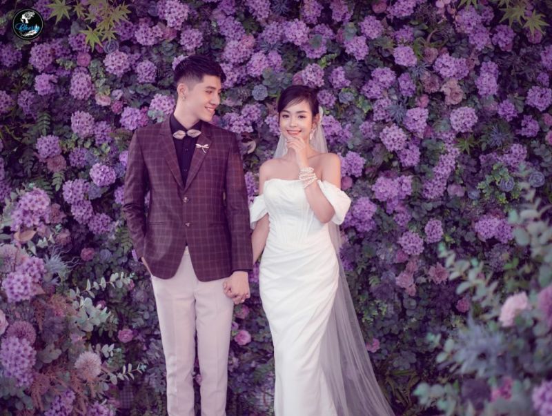 cô dâu và chú rể đứng chụp hình ở tường gắn đầy hoa tím