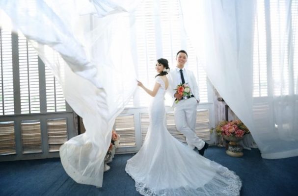 cô dâu và chú rể chụp hình cạnh cửa sổ có tấm èm bay phấp phới màu trắng tinh